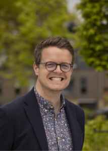 Smiling image of a smartly dressed man with glasses outdoors with trees visible behind him.