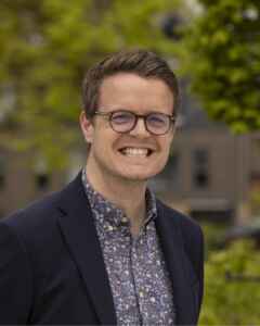 Smiling image of a smartly dressed man with glasses outdoors with trees visible behind him.